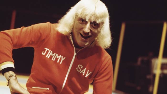 A picture of Jimmy Savile smiling towards the camera and wearing a cardigan that has his name across the front