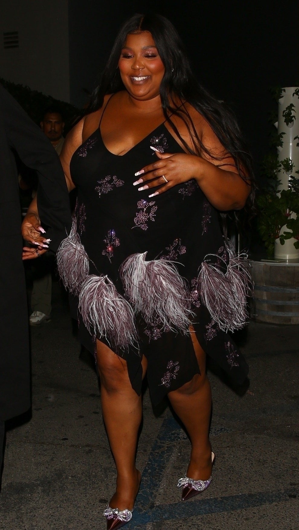 Lizzo wears a short black thin strapped dress with bunches of purple feathers attached to the bottom
