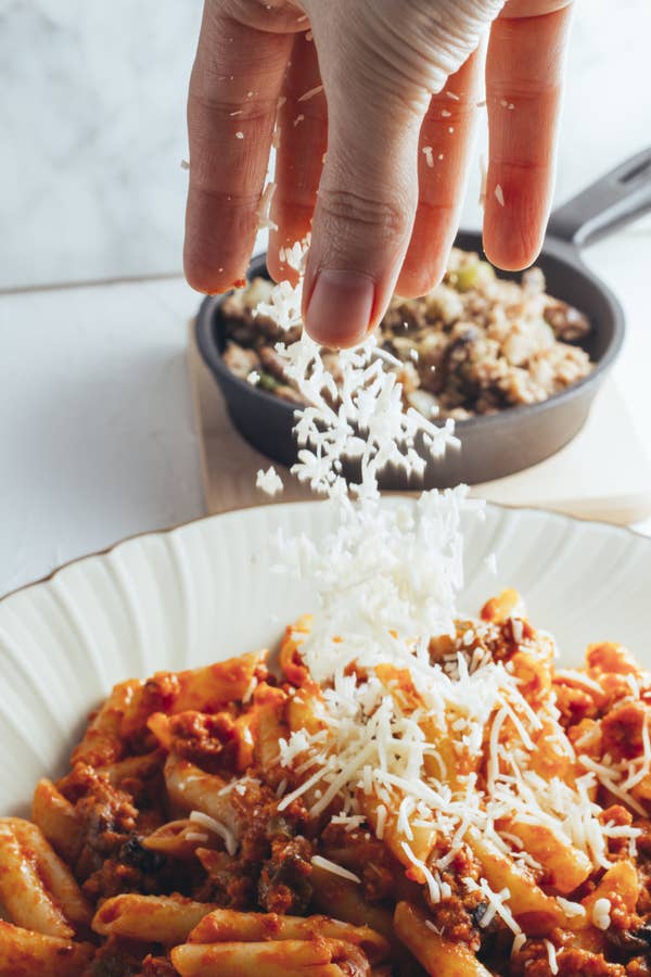 The hand of a woman sprinkling shredded cheese on pasta with tomato sauce