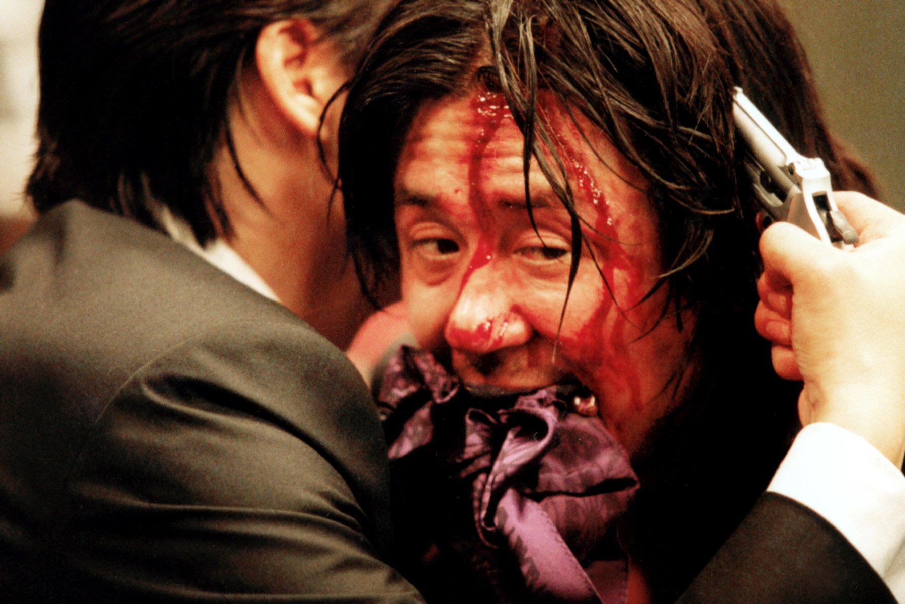 Min-sik&#x27;s character covered in blood while another character puts a gun to his head