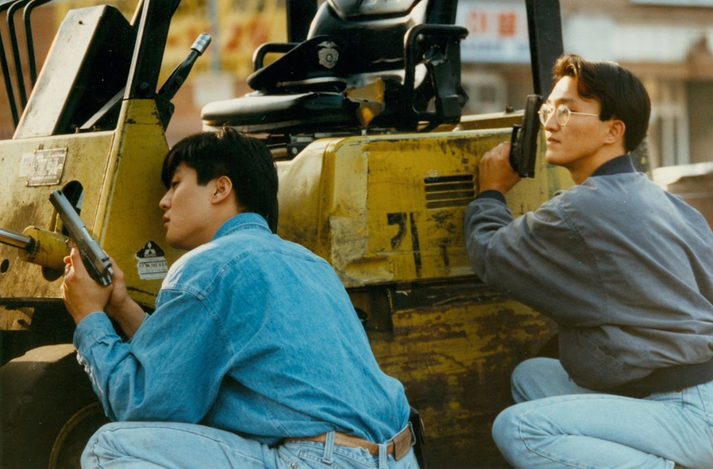 Two Asian American men with guns couch behind a piece of heavy machinery