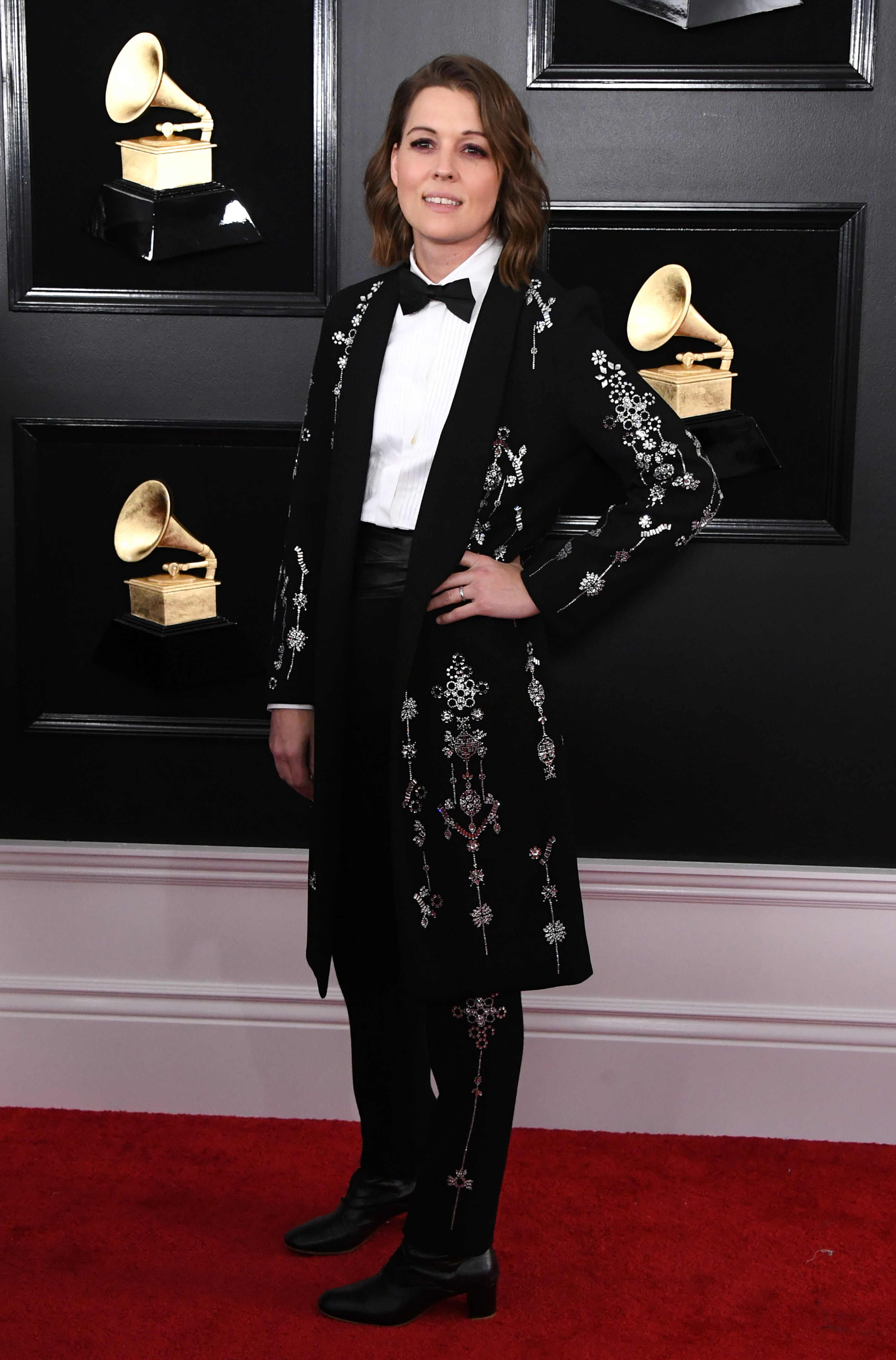 Brandi Carlile at the 61st Annual GRAMMY Awards. She wears a long suit jacket to her knees, which is embellished with silver beaded designs. This continues down the side of her trousers, which she wears with a white shirt, black bow tie and black boots.