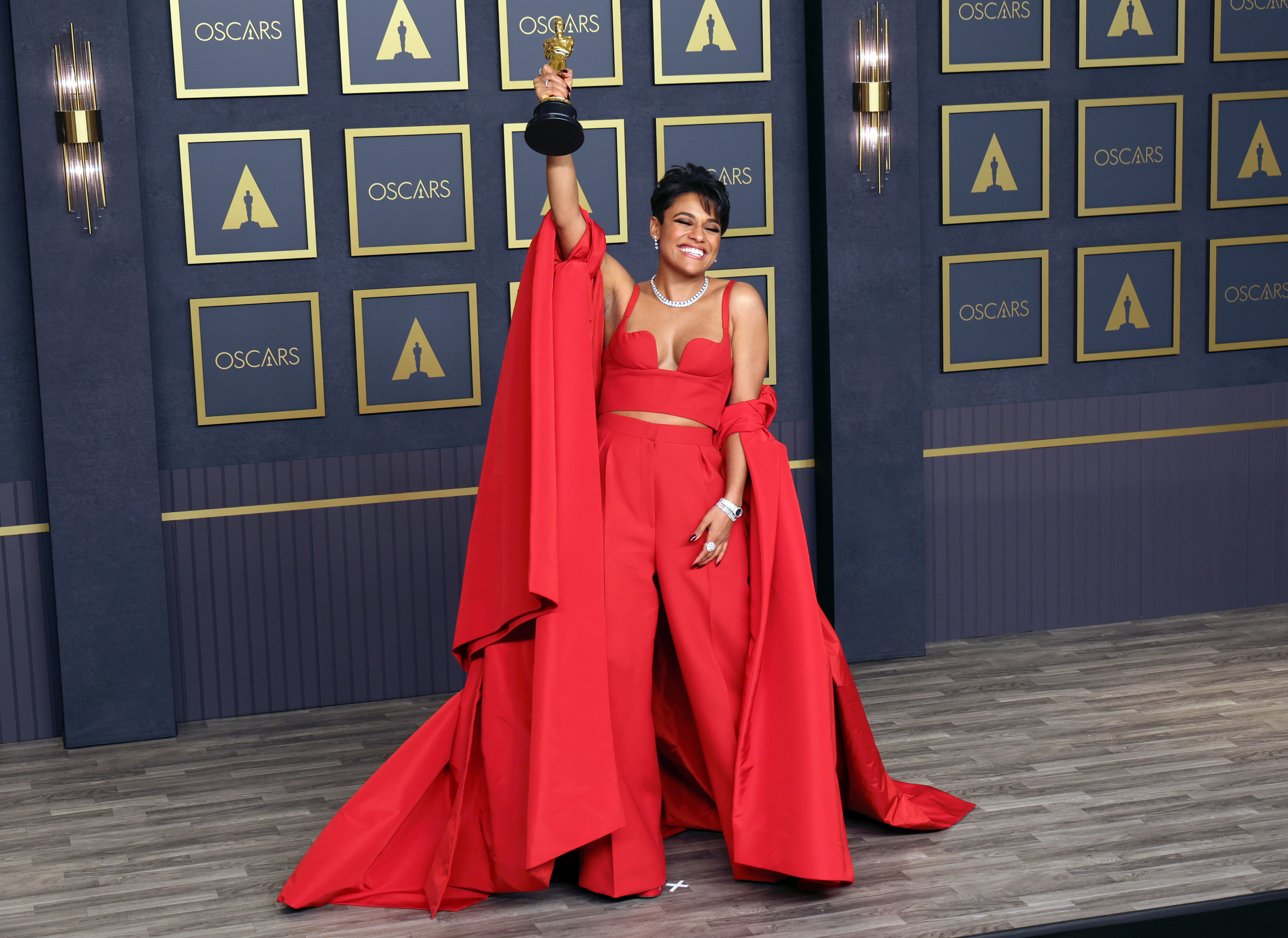 Ariana DeBose poses in the press room at the 94th Annual Academy Awards at Hollywood and Highland on March 27, 2022 in Hollywood, California. She wears a red pantsuit with a vest top and swooping red cape.