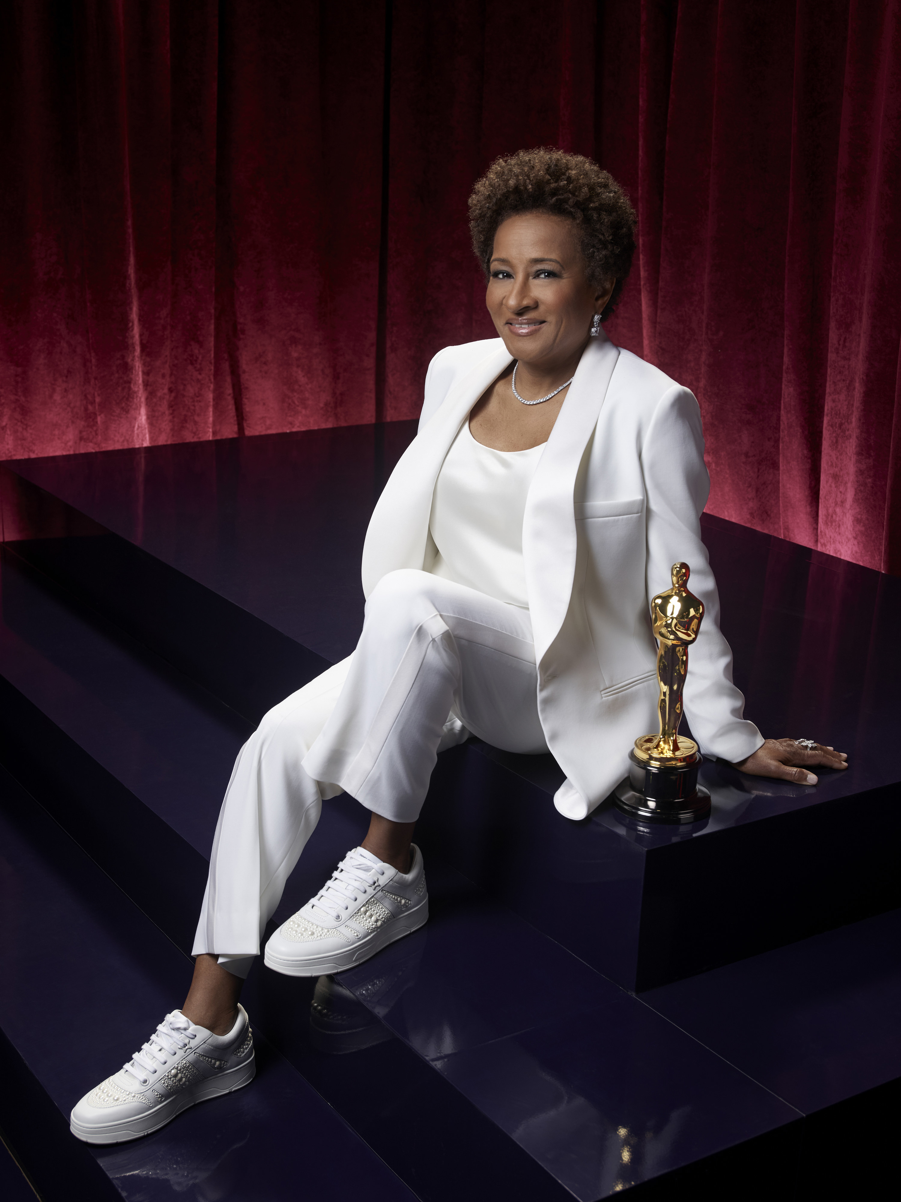 Wanda sits on some black steps wearing an all-white suit with white sneakers and a silver necklace.