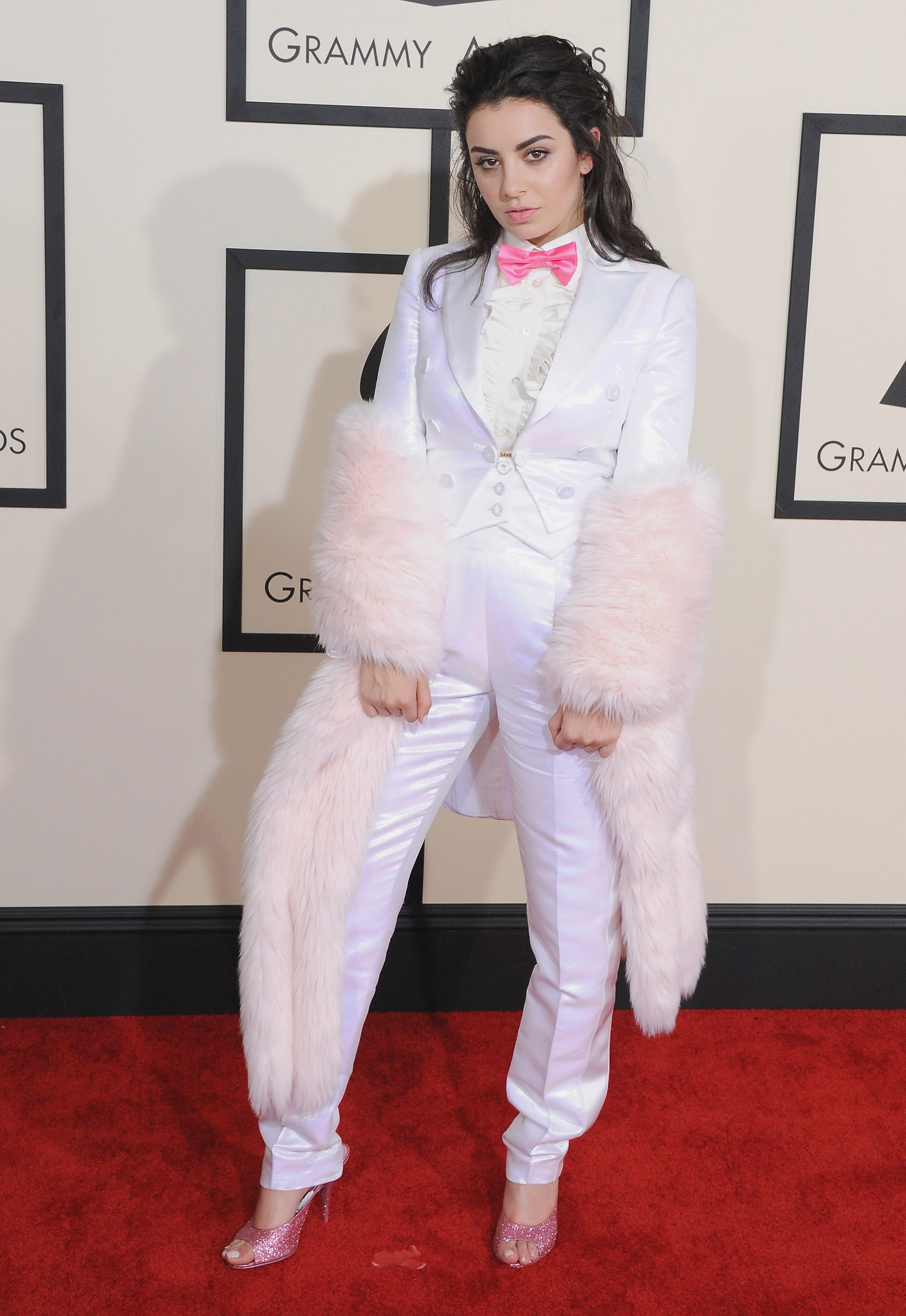 Singer Charli XCX arrives at the 57th GRAMMY Awards at Staples Center. She wears a white tuxedo with frilled chest detail, a pink bow tie, pink peeptoe heels and a white furry coat.