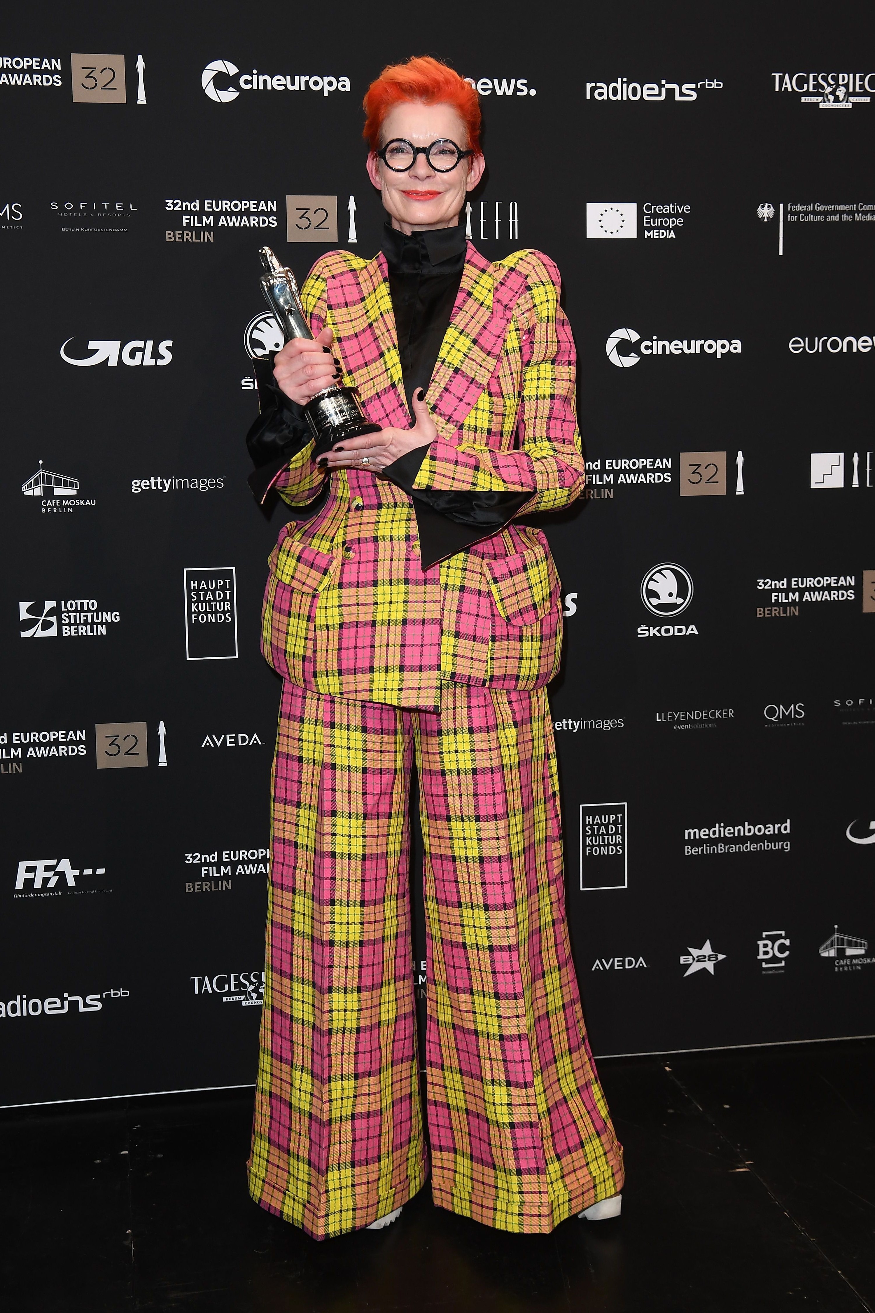 Sandy wears a suit in bright pink and neon yellow plaid, which curves out at the waist. She wears baggy flared trousers in the same pattern.