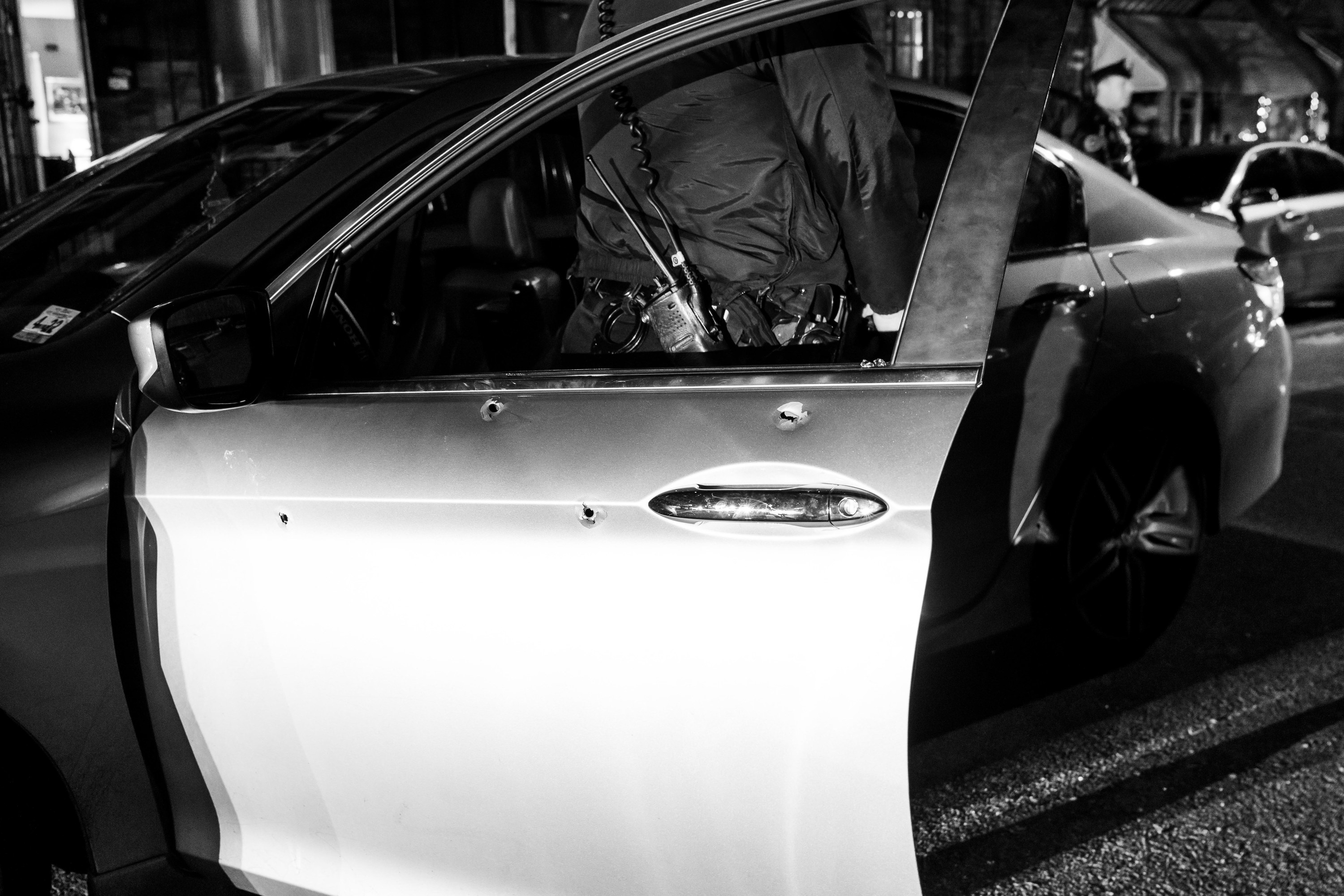 A cop stands behind a car door with visible bullet holes