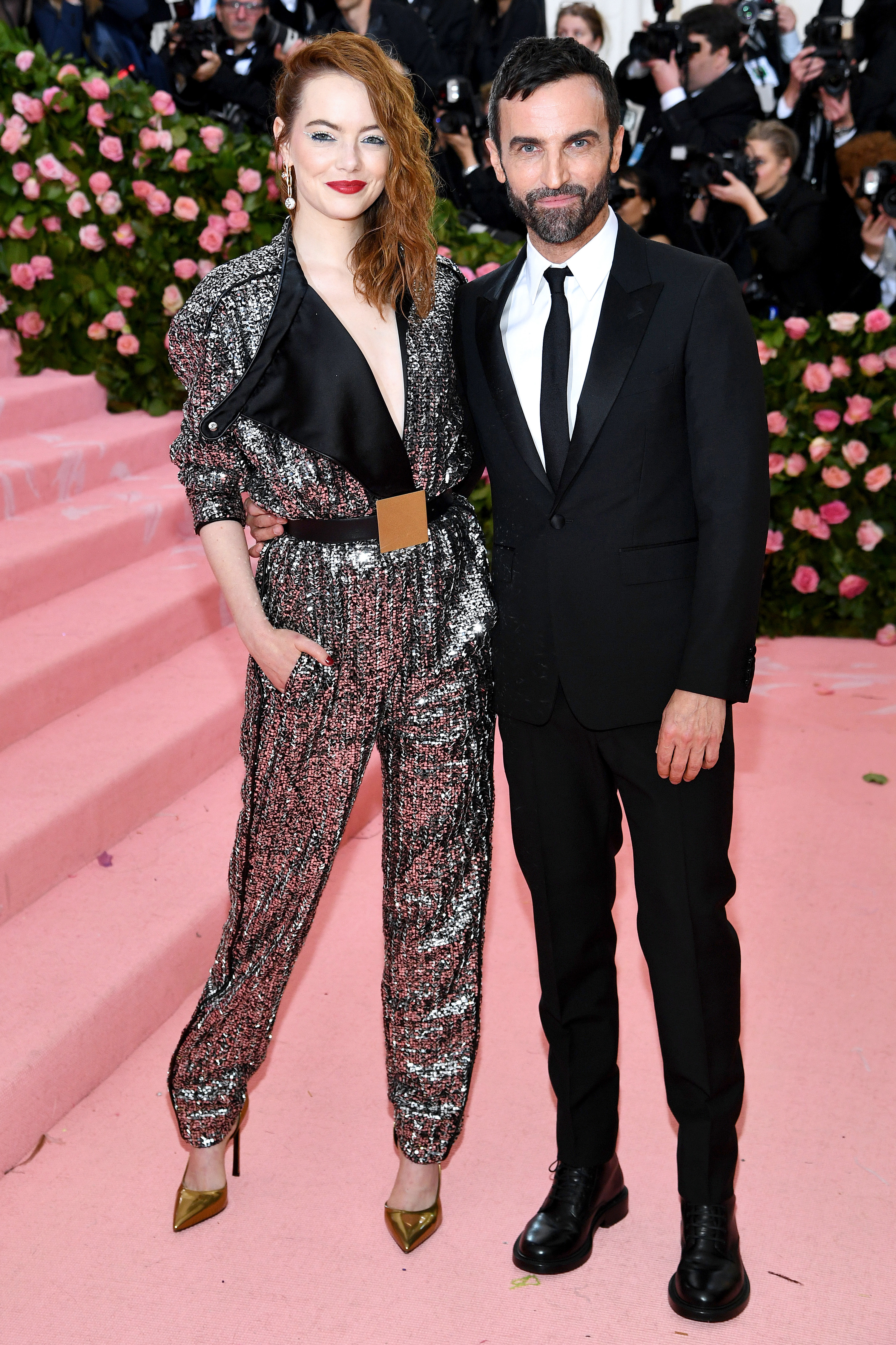 Emma Stone attends The 2019 Met Gala. She wears a metallic, silver, sequinned jumpsuit with a plunging neckline and a black asymmetrical lapel on one side. She teams it with gold pointy toed shoes and a gold buckled belt.