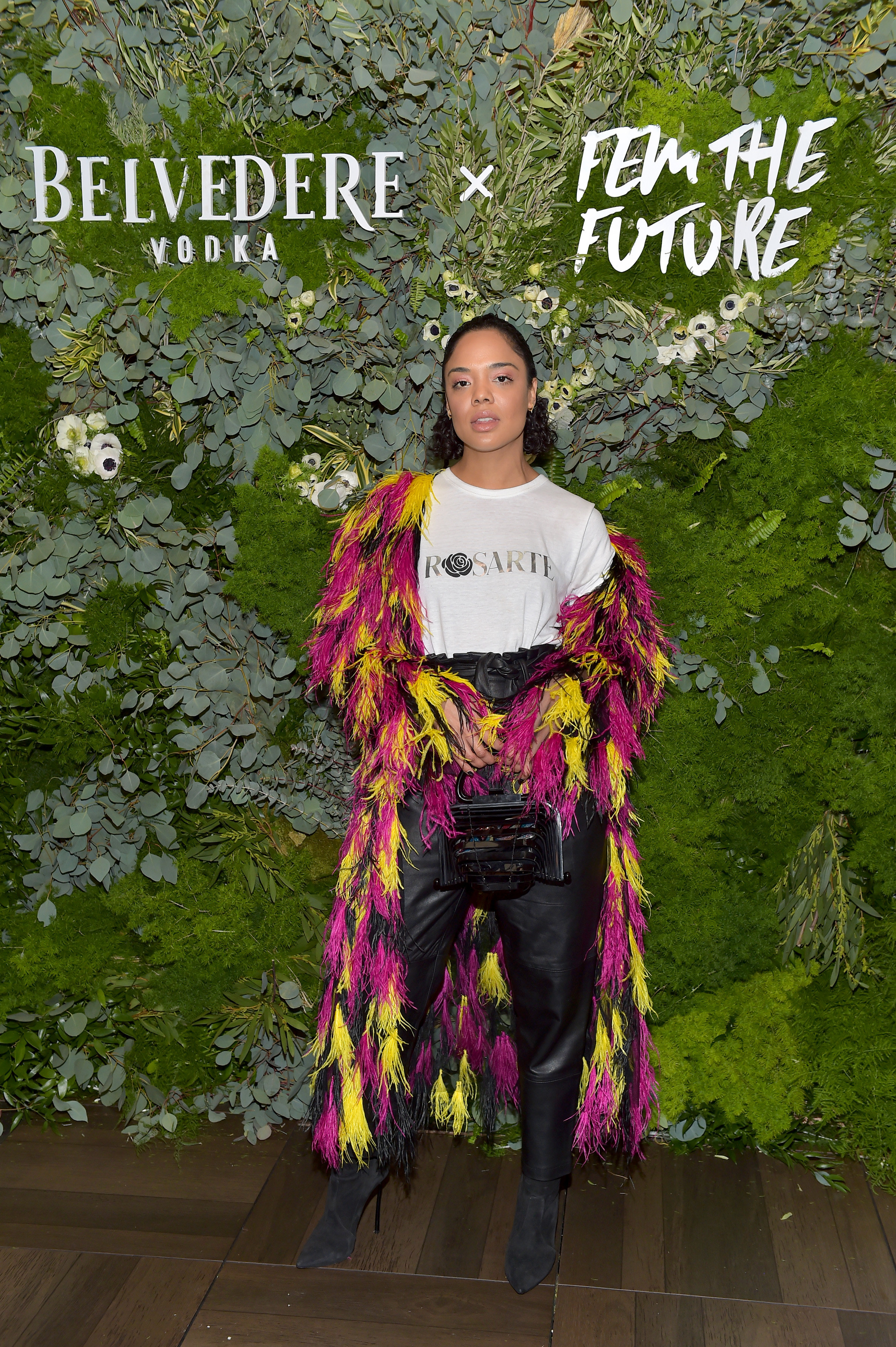 Actor Tessa Thompson attends Belvedere Vodka kick-off &quot;A Beautiful Future&quot; Campaign. She wears black leather trousers, black suede boots, a white t-shirt that says &quot;Rosarte&quot; on it, and a long feathered jacket in black, yellow and fuchsia.