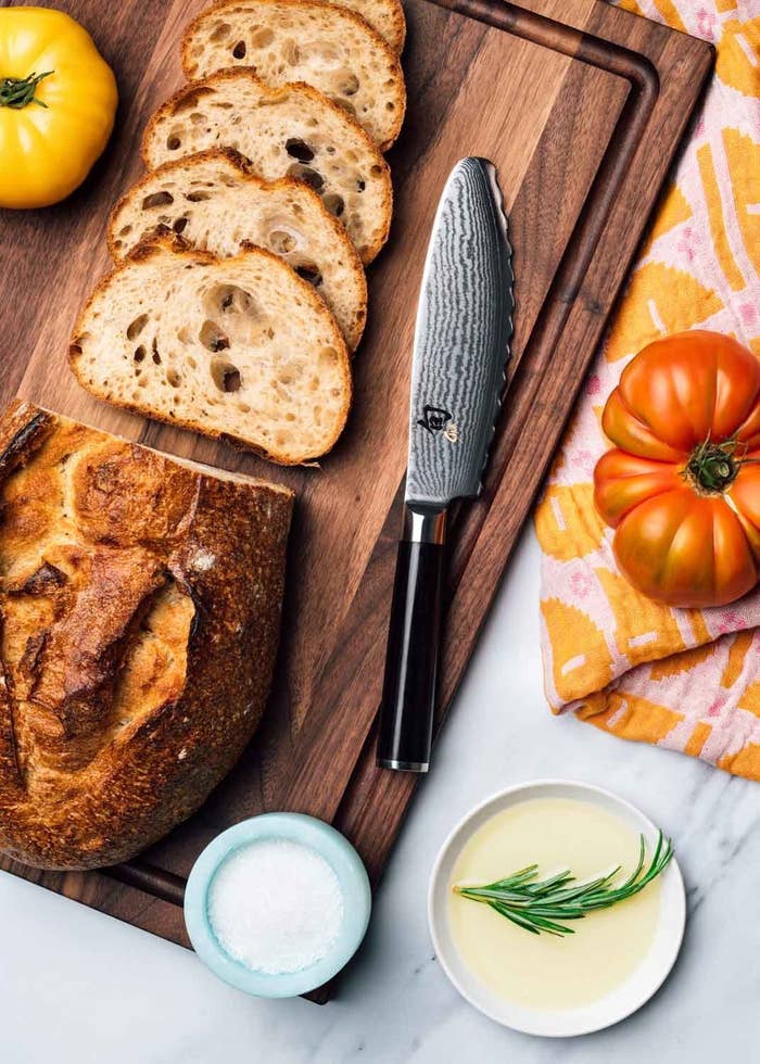 Bread knife on a wooden cutting board, next to sliced bread and a tomato