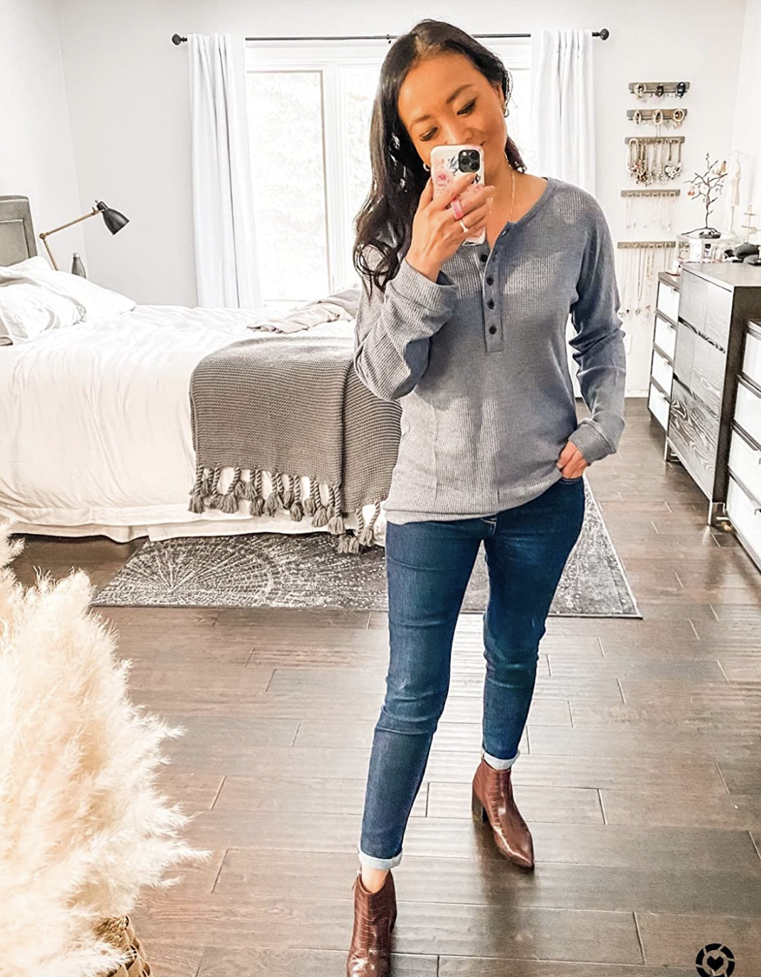 reviewer in a casual buttoned top and jeans, taking a selfie in a mirror in a well-lit bedroom