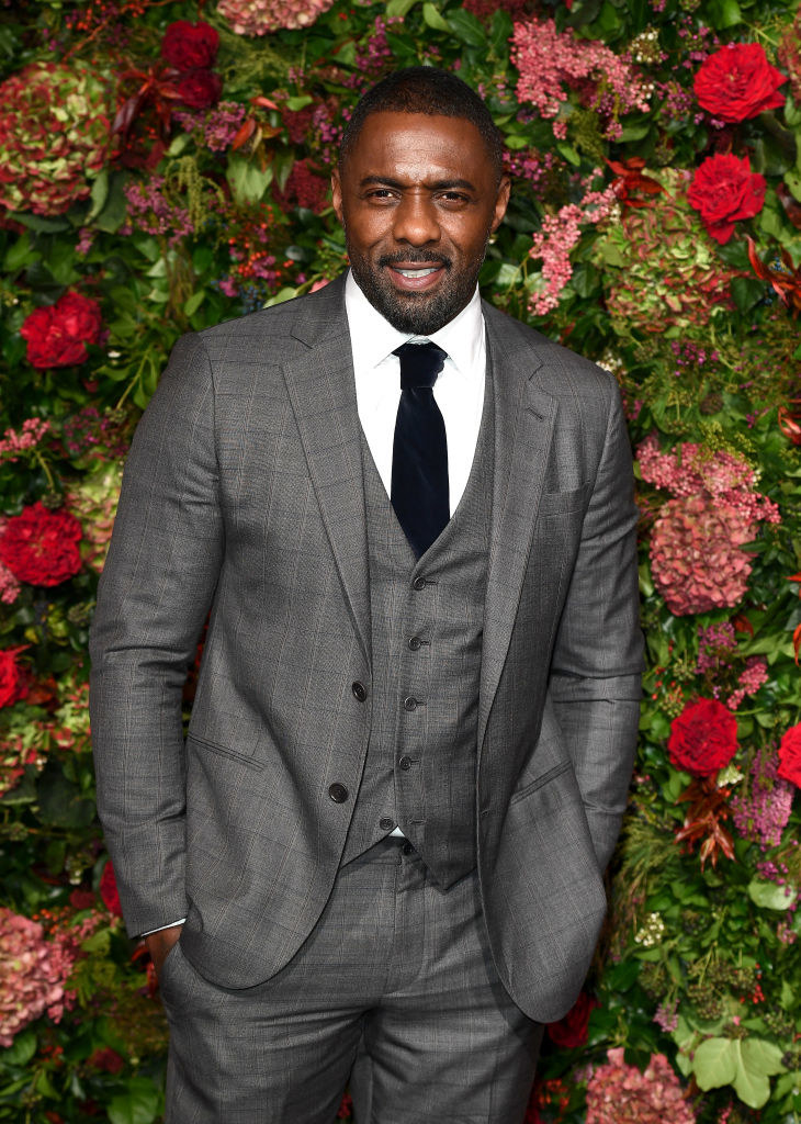 Idris in a three-piece suit against a flowery backdrop with a mustache and slight beard