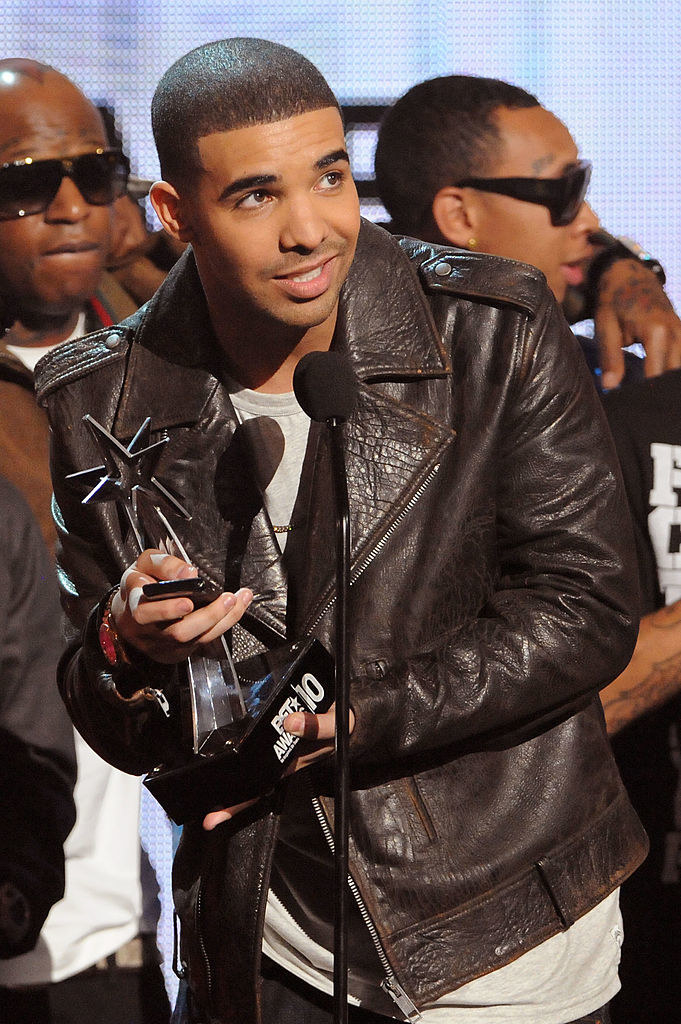 Buzz-cut Drake holding an award at a microphone, light mustache, no beard