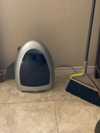 the gray and black bagless, touchless vacuum on a tiled floor next to broom