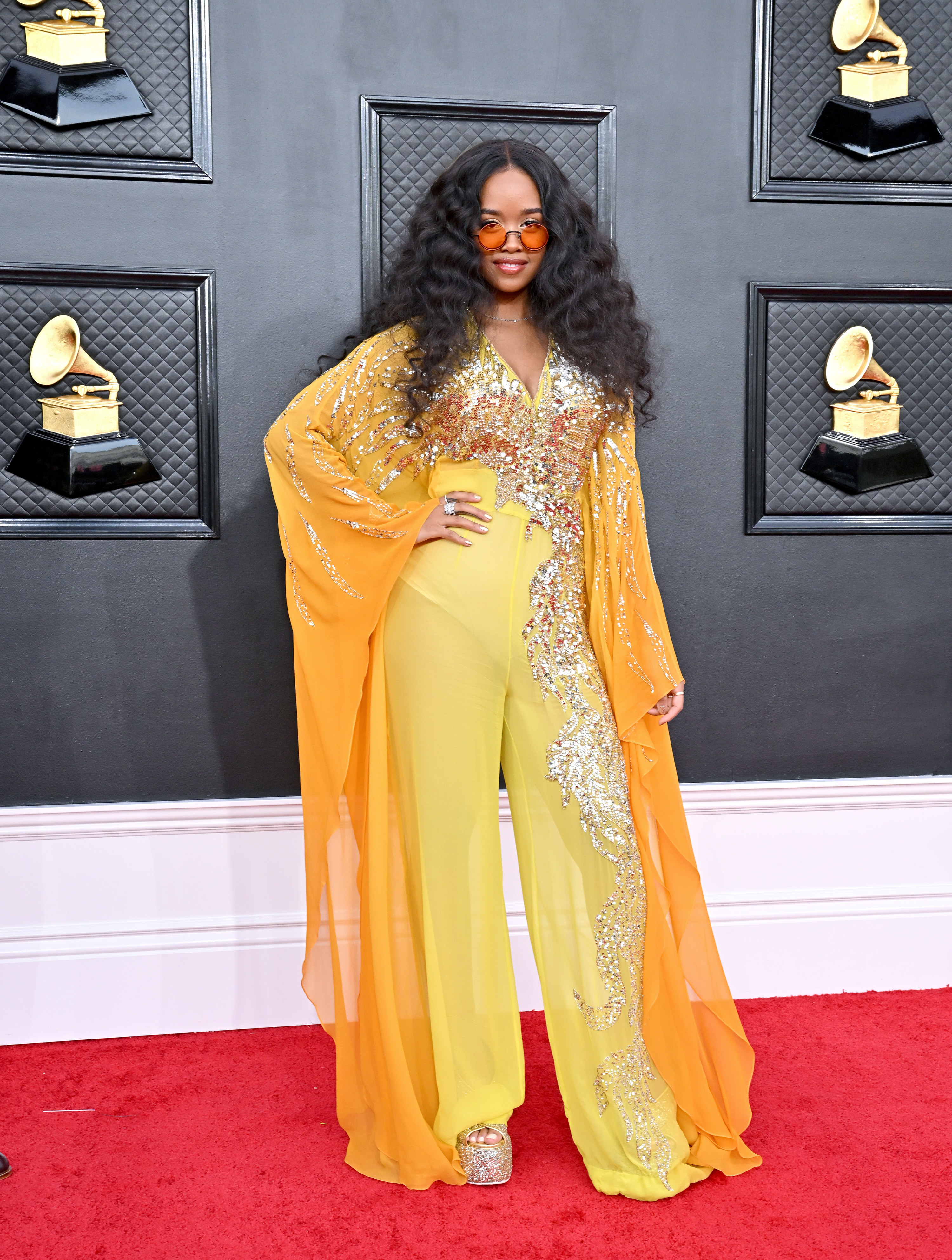 H.E.R. attends the 64th Annual GRAMMY Awards at MGM Grand Garden Arena. She wears a yellow jumpsuit with sequinned waterfall detail from the bust down one leg, with a burnt yellow floaty bolero with sequinned sun rays that reaches to the floor.