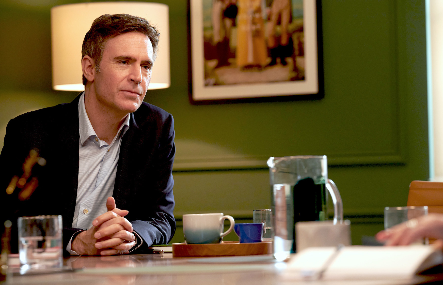 Jack Davenport as Jonathan Nightingale in Ten Percent sits at a desk in an office room and talks to someone off camera