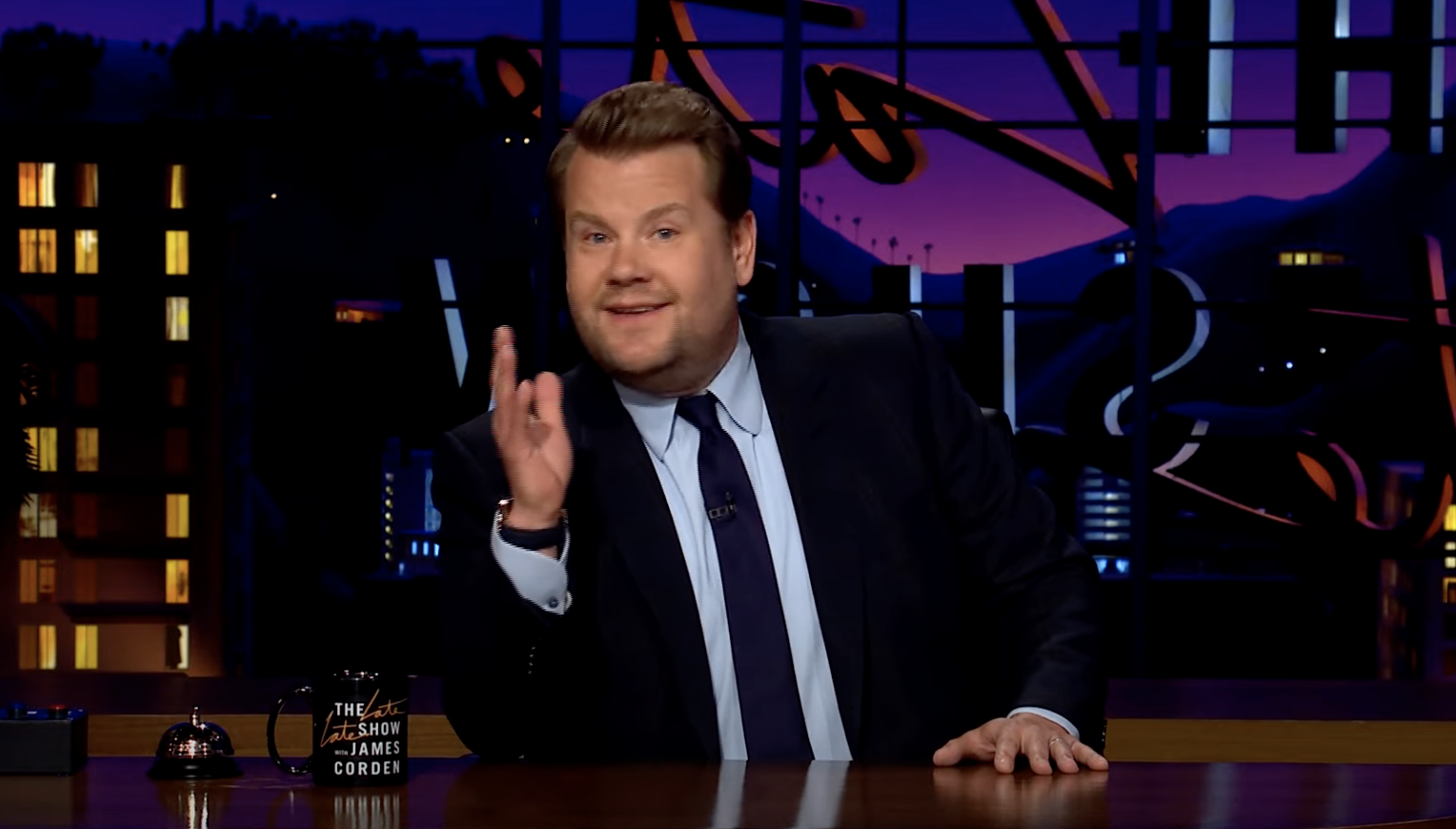 James behind his desk on the set, holding up one hand