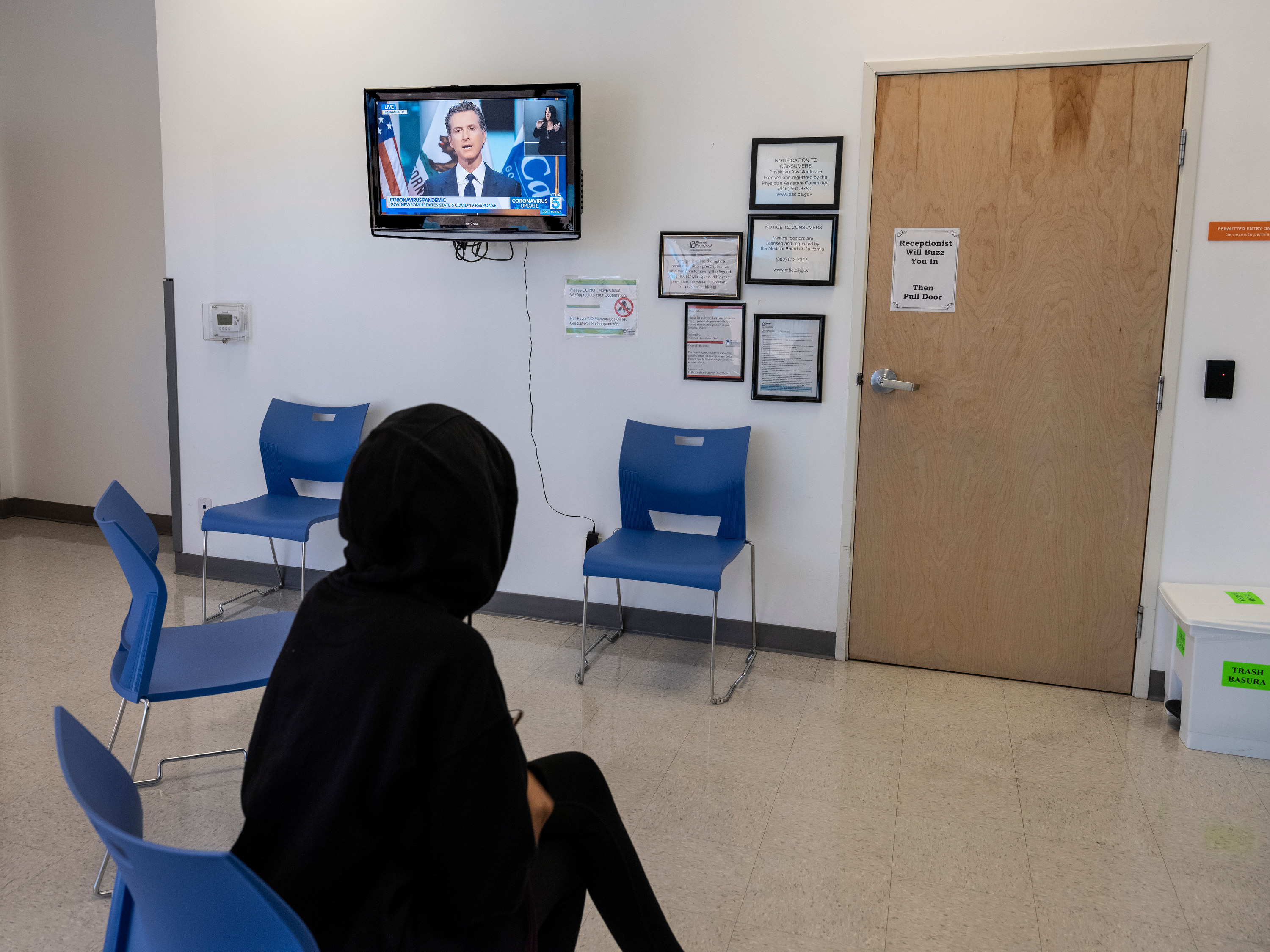A figure in a hoodie sits in a waiting room where a TV is showing news coverage