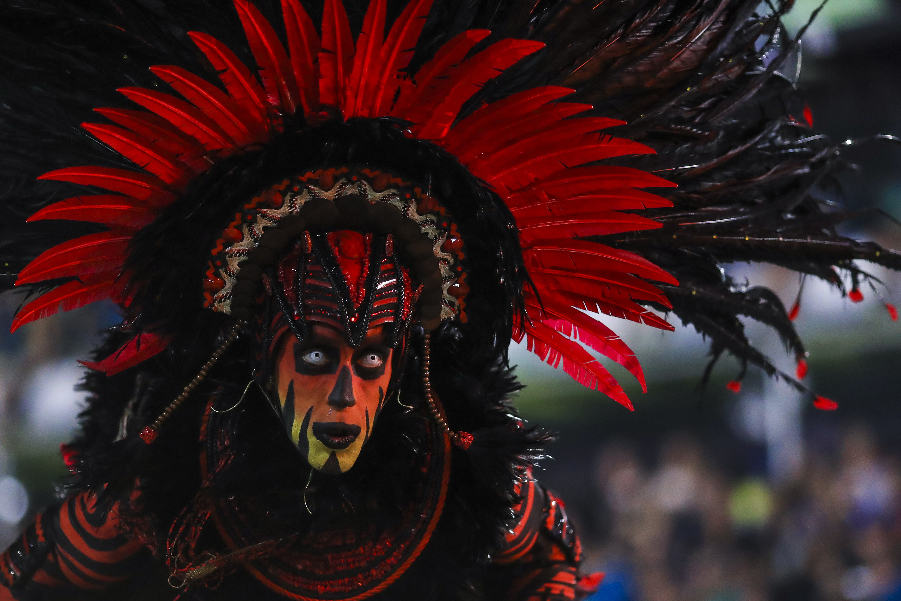 A person wearing face paint, bright contact lenses, and a large feathered headdress