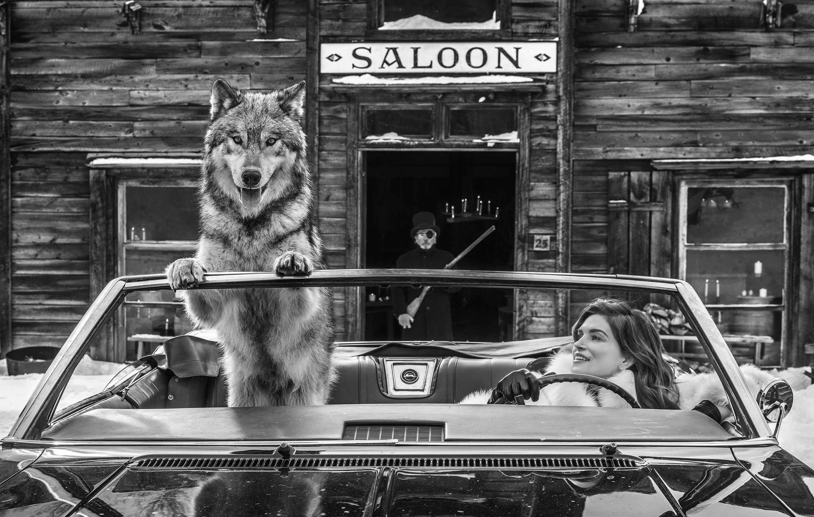 A husky sets its paws on the windshield of a convertible from the passenger seat and Cindy Crawford watches from the driver&#x27;s seat while a man with a shotgun and an eyepatch stands behind them in the doorway of a building marked &quot;Saloon&quot;