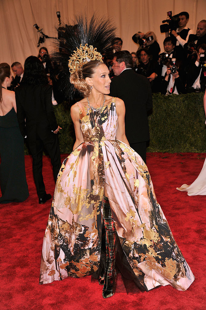 SJP wears a gown with gold foil details and thigh high boots with a large crown with feathers