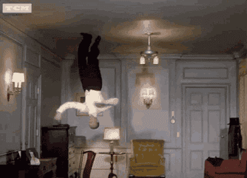 man in button down shirt and slacks tap dances on the ceiling of a living room