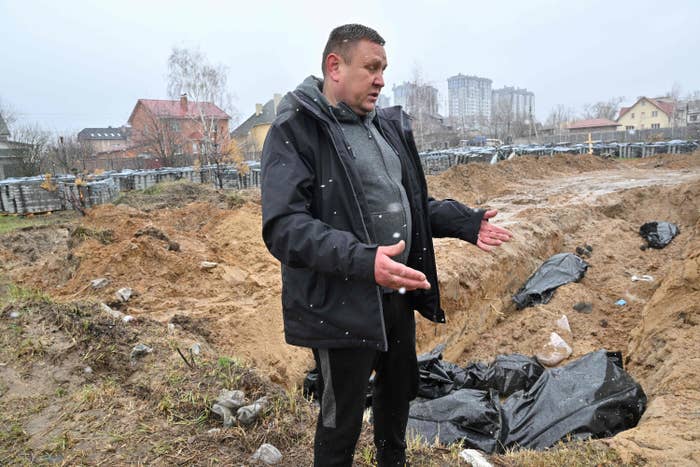 A man stands in front of a large hole in the ground containing bodies in black bags