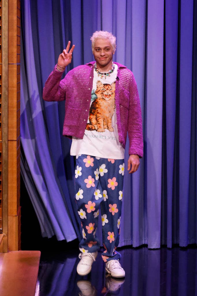 Pete walking on a talk show wearing floral pants, a cat shirt, and pink cardigan