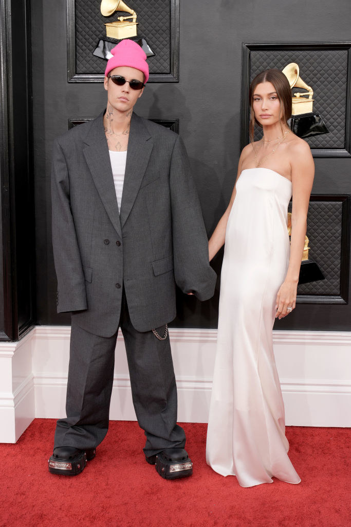 Serious-looking Justin in a boxy suit holding hands with a serious-looking Hailey, in a long, strapless, flowing straight gown
