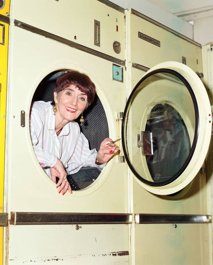 Jude Brown leaning out of a washing machine, holding a cigarette smiling at the camera. She&#x27;s wearing a white shirt with light gray stripes