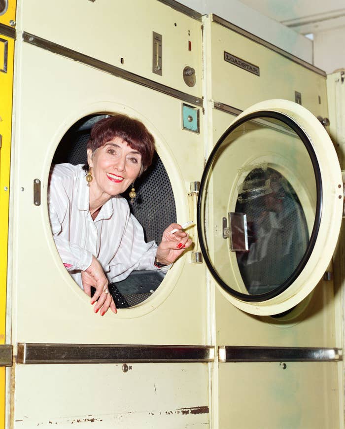 Jude Brown leaning out of a washing machine, holding a cigarette smiling at the camera. She's wearing a white shirt with light gray stripes