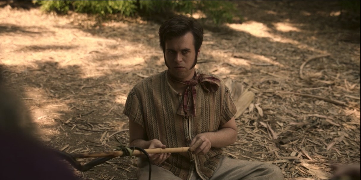 Lucius sits on the ground, holding a stick that he was using to roast a snake over a fire, looking with confusion at his colleagues.