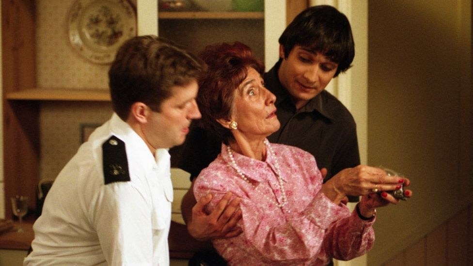 Dot Cotton in a pink floral shirt, leaning backwards on a man in a dark brown top, while he holds her with the help of a police officer