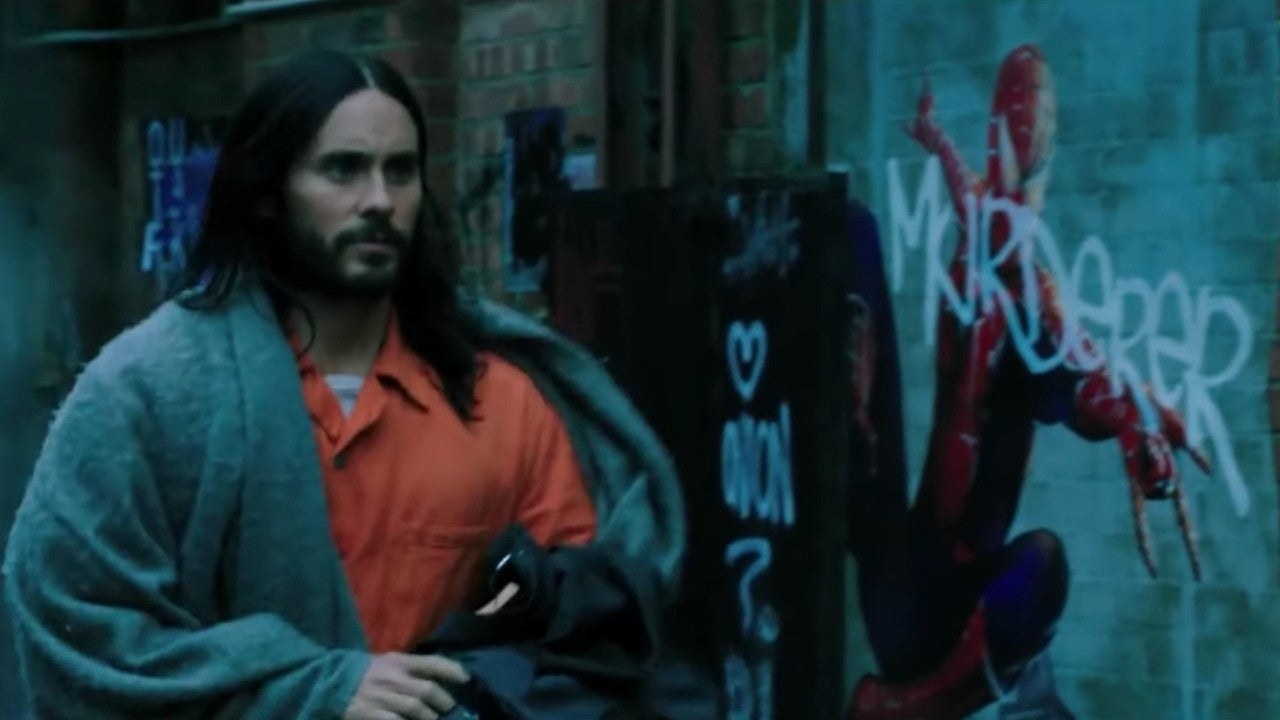 Morbius in an orange jumpsuit with a grey blanket wrapped around his shoulders, walking past some Spider-Man graffiti with MURDERER written across it