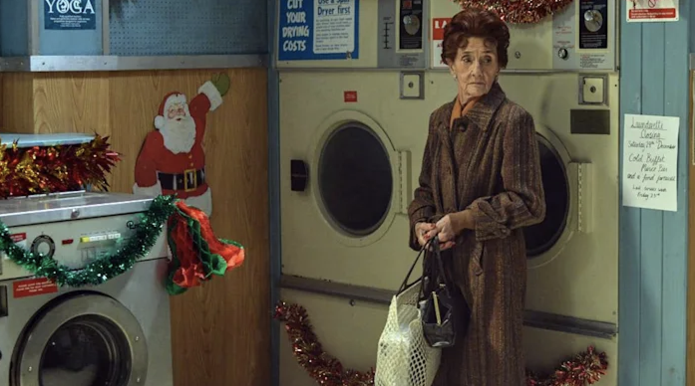 Dot Cotton, standing in front of some washing machines in a laundrette, wearing a long brown coat and holding some bangs. She&#x27;s looking around sadly