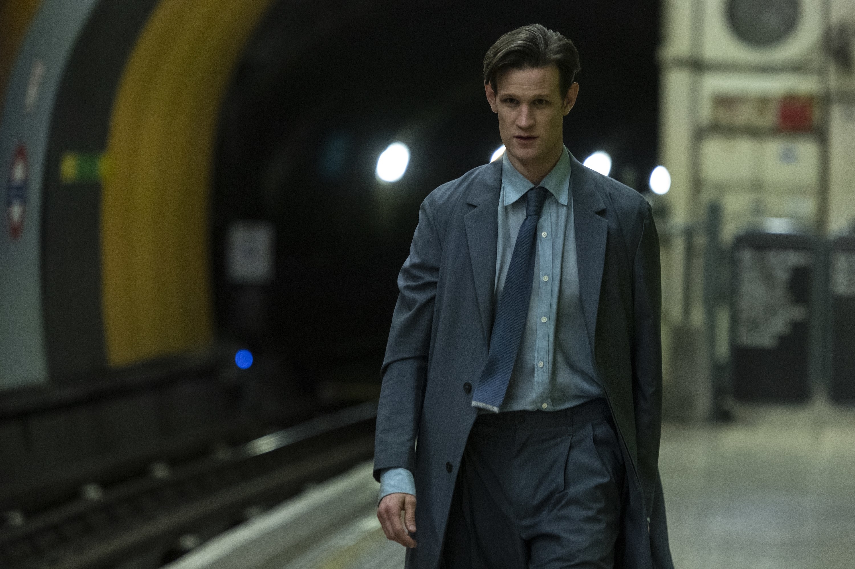 Milo walking down a train platform in a long grey coat, light blue shirt and dark blue tie
