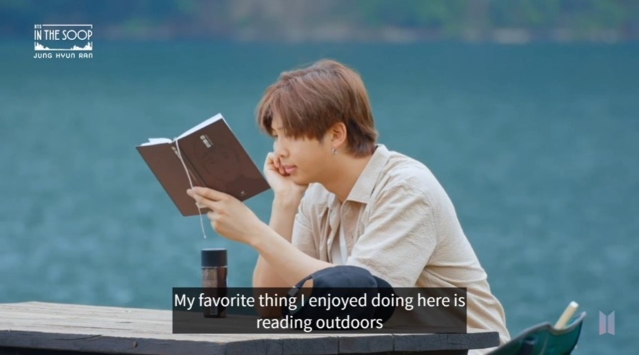 RM sits on a picnic bench by a river, reading with a coffee and a packet of gummy sweets