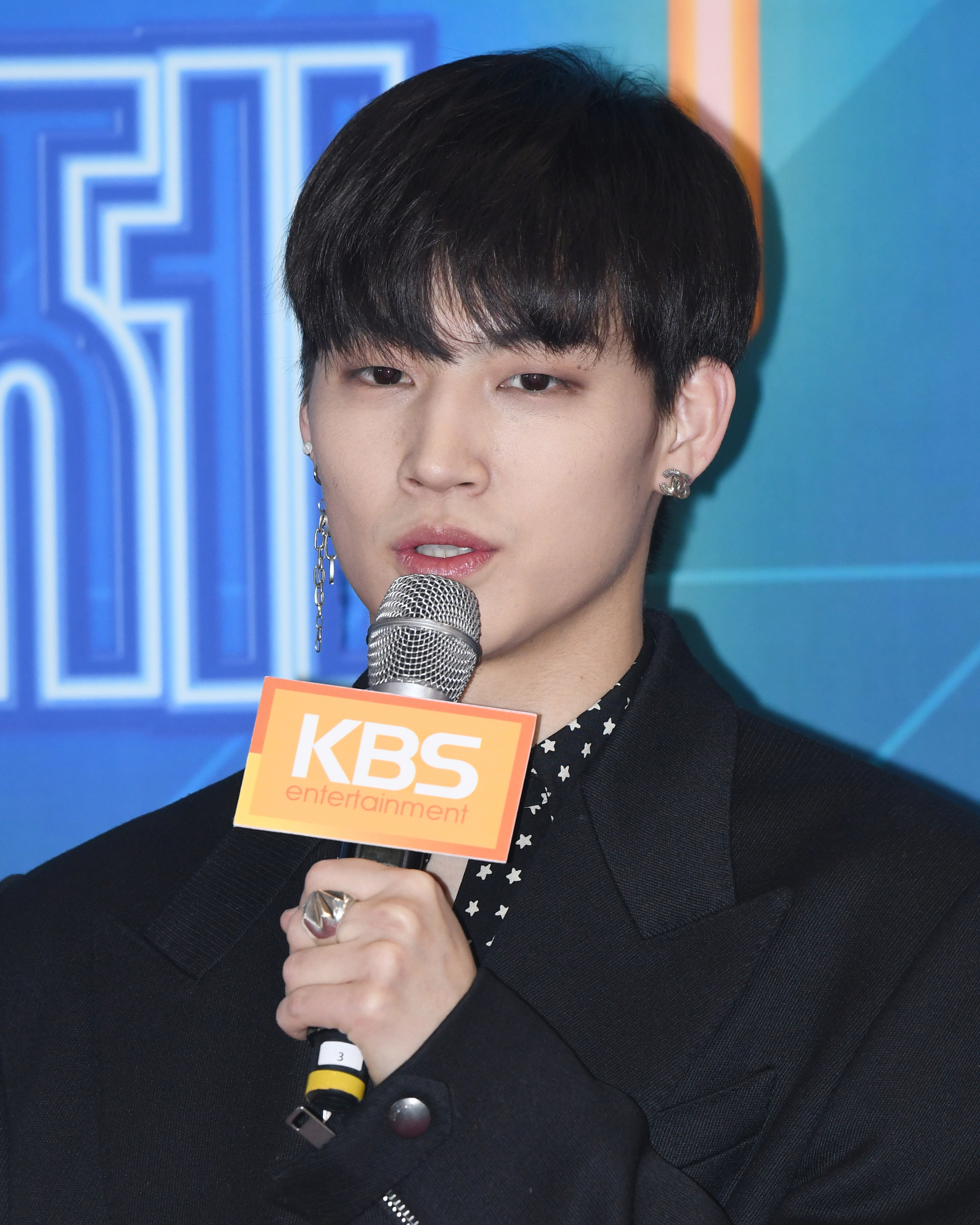 JB holds a microphone to his mouth with a sign on it saying &quot;KBS entertainment&quot;; he has floppy black hair that brushes his eyebrows and wears a long dangling silver earring in one ear