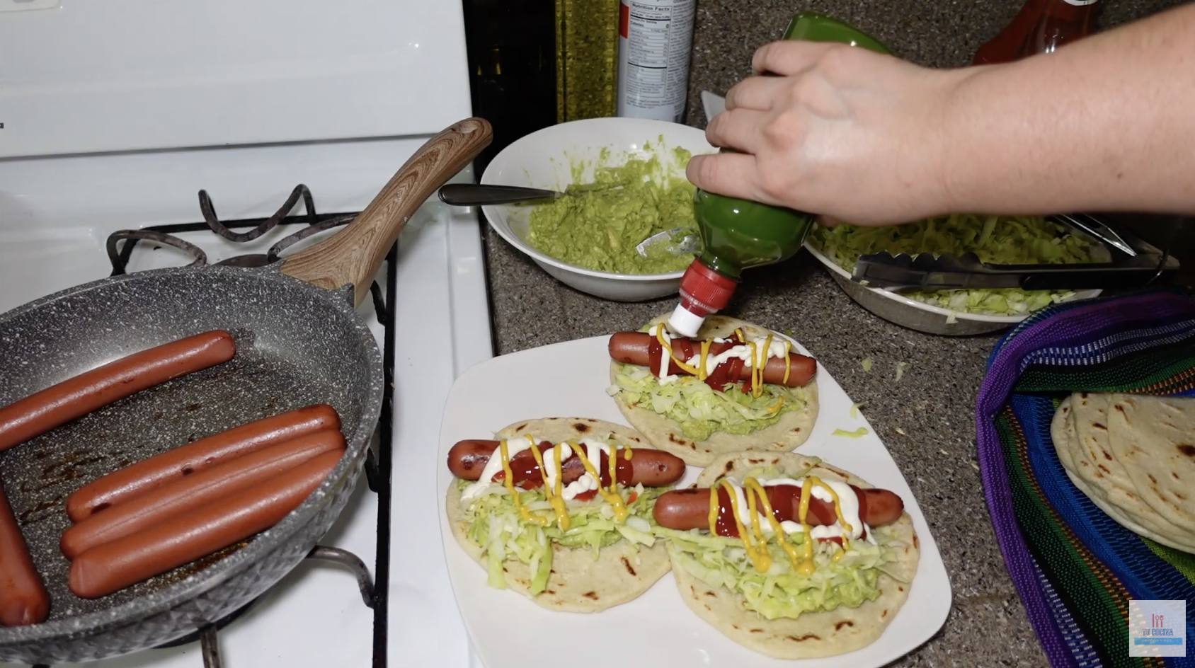 A person adds the condiments to mixtas on a plate as other hot dogs are cooking in a pan