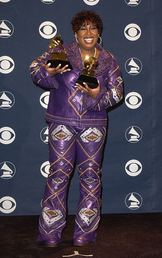Missy smiling and holding two Grammys, wearing a bright-colored, shiny, patterned pantsuit