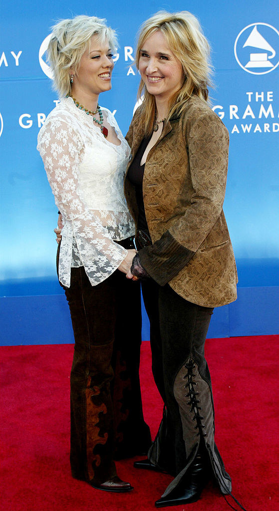 Tammy wearing a long-sleeved, lacy top and pants and holding hands with Melissa, wearing a suede-looking jacket and side-lace-up pants