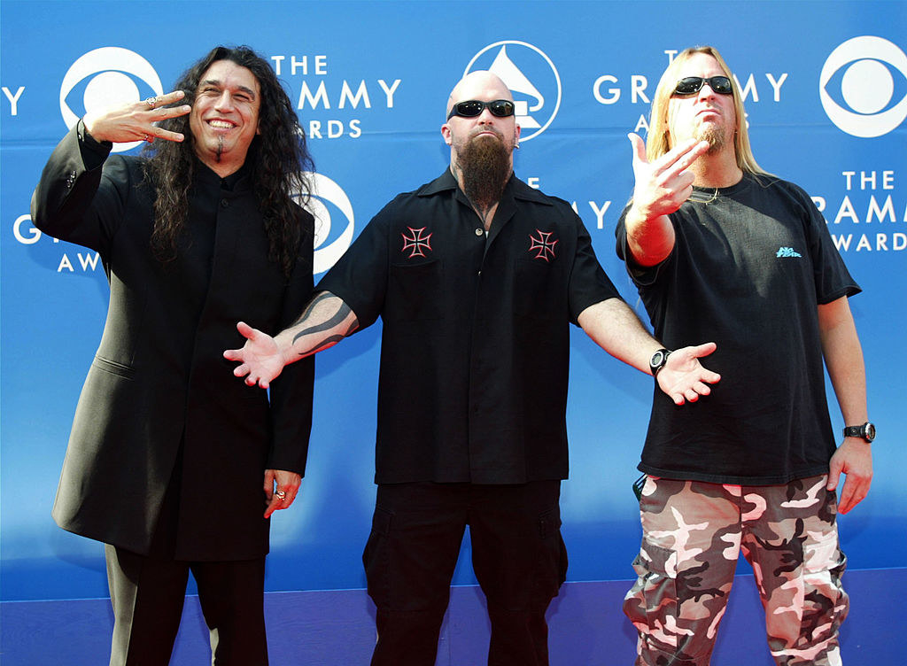 The three members gesturing and wearing a suit, a loose top and loose pants, and a T-shirt and camo pants
