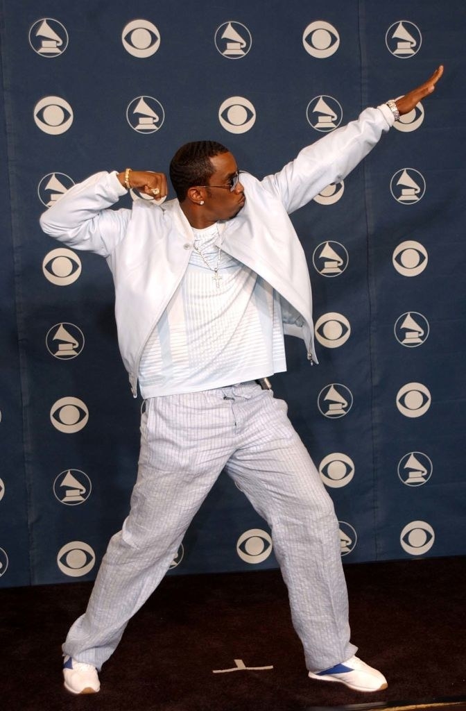 Diddy in a loose, light-colored outfit and striking a pose with one fist raised and the other arm held up