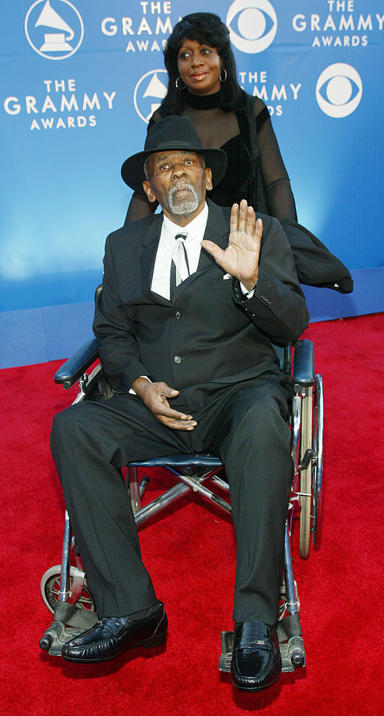 James in a wheelchair and waving and wearing a hat and a suit