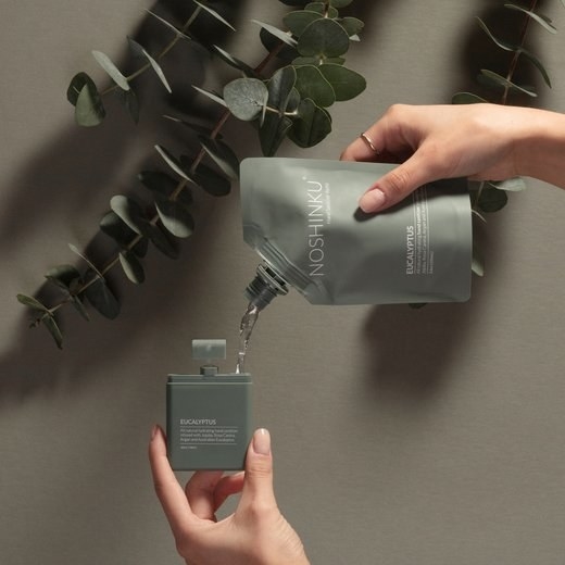 model pouring a sanitizer refill from a bag into a square sage green pocket-sized spray bottle