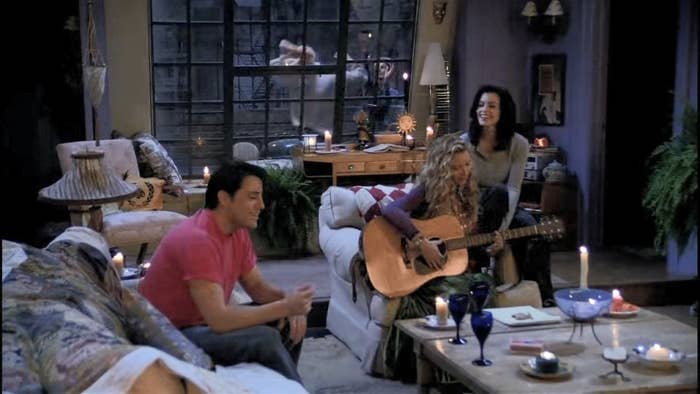 Phoebe Buffay plays the guitar while Monica Geller sits on the chair with her and Joey Tribbiani sits across from them