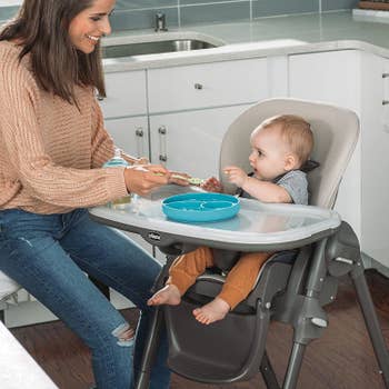 A mother feeding a baby who is sitting in the gray high chair