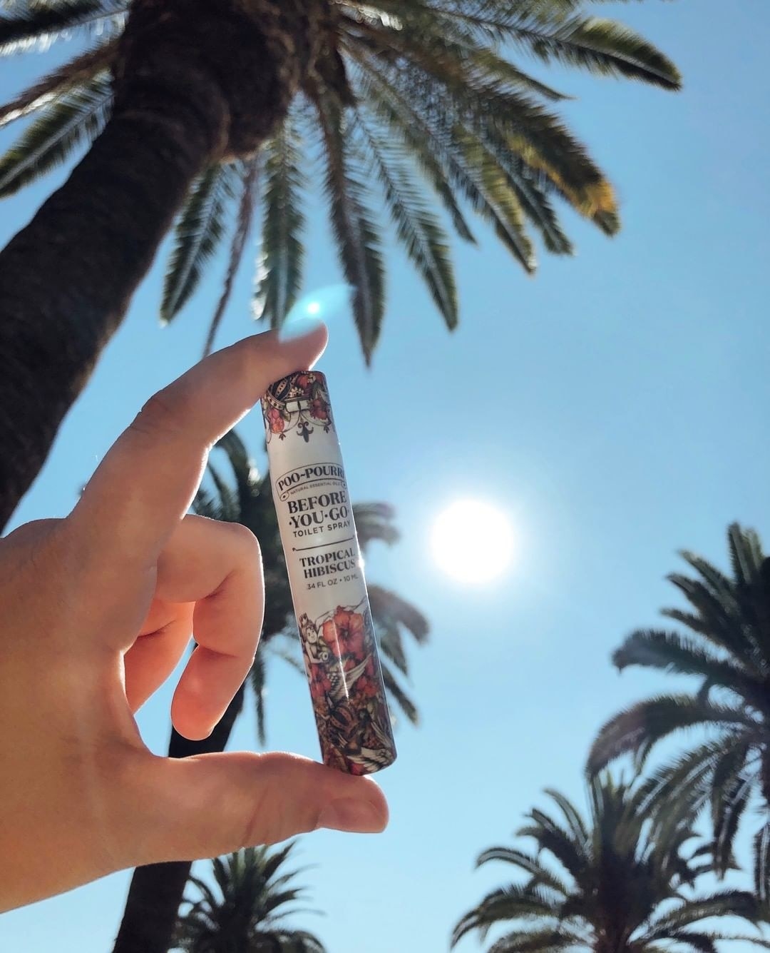 A person holding a tube of Poo-Pourri in front of palm trees