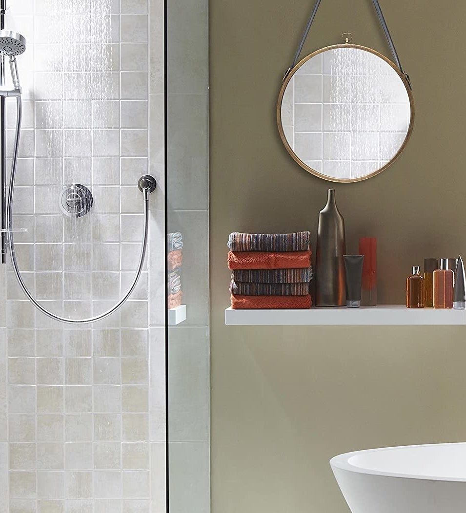 The mirror hanging on a bathroom wall, next to a shower and above a floating shelf with toiletries and towels on it