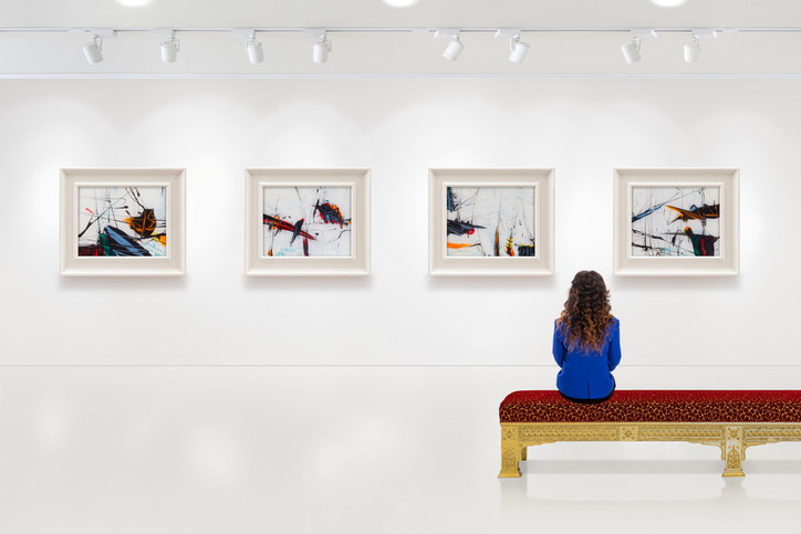 In a exhibition centre, young woman visits an art exhibition and watches artist&#x27;s collection on the wall. Lightened white wall contains four white frames with artist&#x27;s painting.
